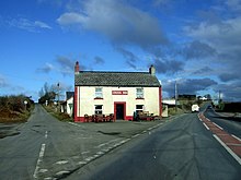 Front elevation of a white-painted two-storey rural pub with a central door, two lower and two upper windows edged with red, situated between two roads under a mostly-blue sky