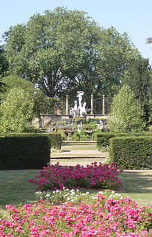 The colour photograph presents a series of lawns leading up to a statue complex. Hedges partially divide these lawns into three sections. The division closest to the camera contains flower beds, while the middle portion holds a circular pond – visible as a stone edge, the far lawn leads up to the statue complex.