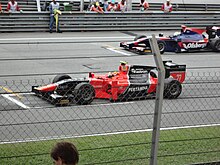 Photographie d'une monoplace de GP2 rouge et noire, vue de profil, derrière un grillage, sur la piste.