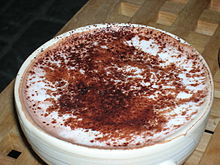 一杯热巧克力，装在厚瓷杯中，杯子盛满了巧克力，表面覆盖着一层泡沫状的奶油，奶油上不均匀地撒了些许可可粉，小部分撒到了杯口上。杯子放置在镂空的木质垫子上。