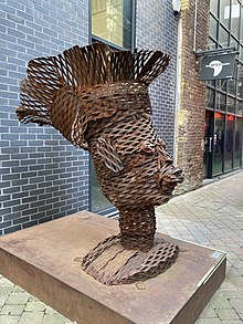 An outdoor scene in Southwark London with a large brown-coloured steel sculpture head in the centre. The steel has a woven effect with exaggerated features and a head covering.