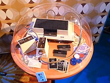 Sous une cloche en verre, figurent la console Odyssey (boite blanche) et certains de ses accessoires (cartes à jouer, dés).