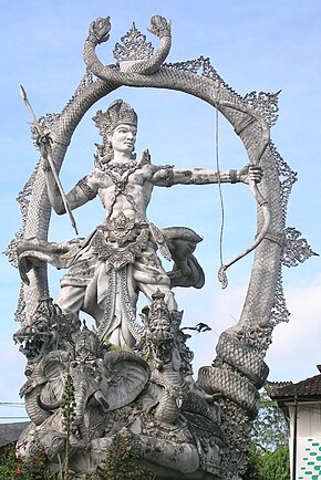 A grey silver statue of Arjuna looking across, surrounded by an oval. She is using a bow and arrow.
