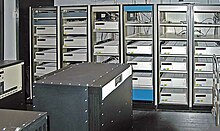 A room with a black box in the foreground and six control cabinets with space for five to six racks each. Most, but not all, of the cabinets are filled with white boxes.