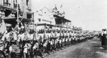 Black soldiers in light-coloured shirts, shorts and hats march down a street of southern African colonial appearance, rifles shouldered