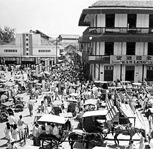 Crowd at a busy street intersection. There are horse-drawn carriages in the foreground while a three-story building (with the sign "Kam Leng") and a single-story building (with the sign "Chunghua Bioscoop") stand in the background on adjacent corners of the intersection.