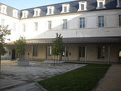 Cour carrée formée par des bâtiments de trois niveaux, aux murs blancs et à toit d'ardoise noire.