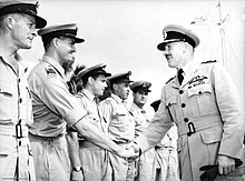 Deux hommes en uniformes militaires clairs avec des casquettes à visière se serrent la main devant une rangée d'hommes habillés de la même façon.