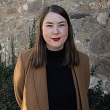 Foto einer Frau, die mit schwarzen Rollkragenpullover und braunem Mantel vor einer Steinmauer steht