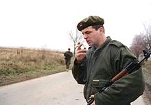 A young man wearing battledress and a beret with a Serbian flag badge stands on a road, smoking a cigarette and holding an AK-47 rifle in the crook of his arm.