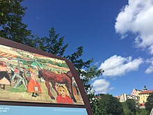 Eine Metallstele im Anschnitt, darauf ein farbenfrohes Bild von Paula Wimmer, das das Treiben auf dem Dachauer Volksfest zeigt. Dahinter sieht man die Altstadt von Dachau auf dem Berg.