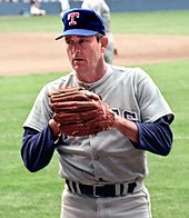 A man in a gray baseball jersey wearing a dark blue baseball cap with a "T" at the centre and a brown Rawlings on his left hand prepares to pitch with the windup.