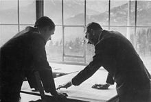 Photo noir et blanc. Adolf Hitler (à droite) et Albert Speer (à gauche), tous deux de profil, en costume sombre, discutent, la tête penchée au-dessus d’une carte étalée sur une plan. En arrière-plan, on voit, à travers une baie vitrée, un paysage de montagne.