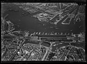 Luchtfoto van het IJ en omgeving, gezien in noordelijke richting. Op de voorgrond het het Damrak en het Stationseiland, op de achtergrond Amsterdam-Noord; circa 1930. Luchtvaartafdeeling, 1920-1940.