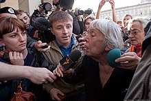 Une femme âgée entourée d'une foule brandissant des micros en sa direction.