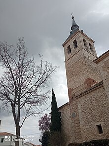 Torre de la iglesia parroquial de Torres de la Alameda, Asunción de Nuestra Señora. Es una torre alta con campanario y rematada en un chapitel. En el lado izquierdo de la imagen hay un árbol.