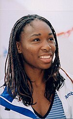 A black woman is serving the ball, and is wearing a white sleeveless top and blue skirt