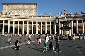 Farbfotografie von einem Platz mit Säulengang und Figuren darauf. Ein Springbrunnen ist auf der rechten Seite zu sehen und einige Menschen sind im Vordergrund. Hinter einer Absperrung am Säulengang hat sich eine lange Menschenschlange gesammelt. Über dem Säulengang ist links der obere Bereich eines Gebäudes mit vielen Fenstern zu sehen.