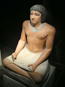 黒髪の、恐らくは中年男性があぐらをかいている写実的な彩色石像。パピルスの巻物の読み物を石彫で模したものを膝に載せている