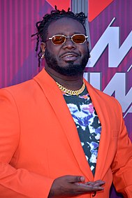 T-Pain steht vor einer lila Wand mit weißer Aufschrift, die verdeckt ist. Er trägt ein orangenes Sakko, ein schwarzes Hemd mit Blumenaufdruck, eine goldfarbene Halskette, einen Dreitagebart, eine Sonnenbrille mit roten Gläsern und die schwarzen Haare als kurze Dreadlocks.