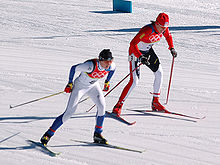 Photographie de deux fondeuses en course, bustes relevés et bâtons en arrière, l'une en combinaison blanche devançant l'autre en combinaison rouge.