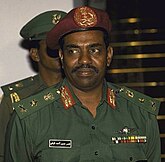 This is a coloured photograph of President Omar al-Bashir in 1990 in military attire, wearing a green uniform adorned with insignias, ribbons, and rank decorations. He is also wearing a maroon beret with an emblem at the centre. The individual has a composed yet stern expression, with a mustache and a focused gaze slightly off-camera. In the background, another person in a similar green uniform is partially visible, contributing to the formal and official atmosphere of the scene.