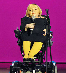 Stella Young sitzt in ihrem elektrischen Rollstuhl, trägt eine senfgelbe Strumpfhose und ein schwarzes Kleid. Sie hat blondes halblanges Haar und hält ein Mikrofon. Der Hintergrund ist pinkfarben.