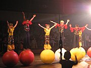 Five children in bright red and yellow costumes balance atop red and yellow spheres.