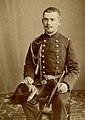 Photographie en noir et blanc d'un homme assis, portant une tenue de bordache, le poignard à la ceinture et le bicorne à la main.