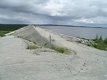 On voit la pointe d'un barrage, une barrière dʼaccès fermée et une grande étendue dʼeau à droite. À l'arrière-plan, la structure de lʼévacuateur de crues sʼélève.