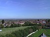 View of the roofs of houses amongst trees and fields. Hills in the distance under a blue cloudless sky.