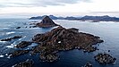 Eine Insel aus schroffem, dunklem Gestein ragt aus dem Meer, darauf ein einzelnes weißes Haus, im Hintergrund eine weitere Insel und das Festland