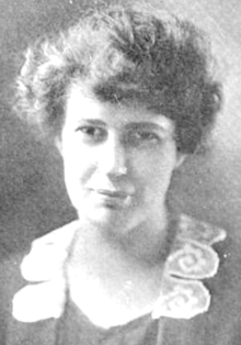 A white woman with hair in a loose updo, wearing a jacket embellished with lace on the lapels