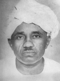 This is a black-and-white portrait of Mahmoud Mohammed Taha wearing a traditional Sudanese turban wrapped neatly around their head. Taha has a calm and serious expression, with a direct gaze at the camera. Taha is dressed in a simple white garment, which blends seamlessly with the soft, light-toned background.