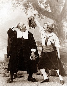 Photo of two white men standing side by side, both amused; the older one is bald and looks up at his wig, caught on a tree; the younger man laughs