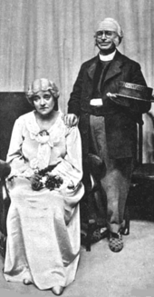 old white woman seated, looking glum, with elderly white clergyman standing to her right