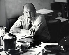 Black and white photograph of Williams in a light colored shirt and dark tie sitting with his crossed arms propped on a desk