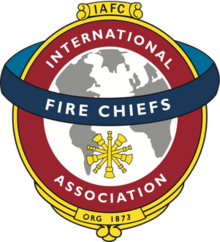 A white and gray globe surrounded by a red circle reading "International Association," with a blue band running horizontally across the circle reading "Fire Chiefs"
