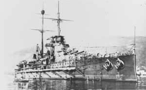 A large battleship sits still in the water near land. Several small boats can be seen in the foreground while a large hill appears behind the battleship in the background.