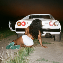Cover art for "Snooze": SZA in white shorts, sitting on the ground in front of the trunk of a white Ferrari 308 GTS.