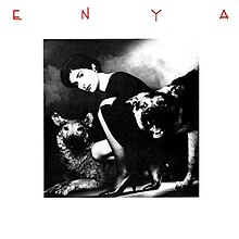 A black and white photograph of a brunette woman with a dark dress that is crouched with two wolves. The image is surrounded by a white border and the word "Enya", stylized in all capital letters, is placed on the top corner of the picture.