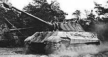 A large tank throws dust from its tracks as it drives by, a man is casually sitting on the front of the turret.