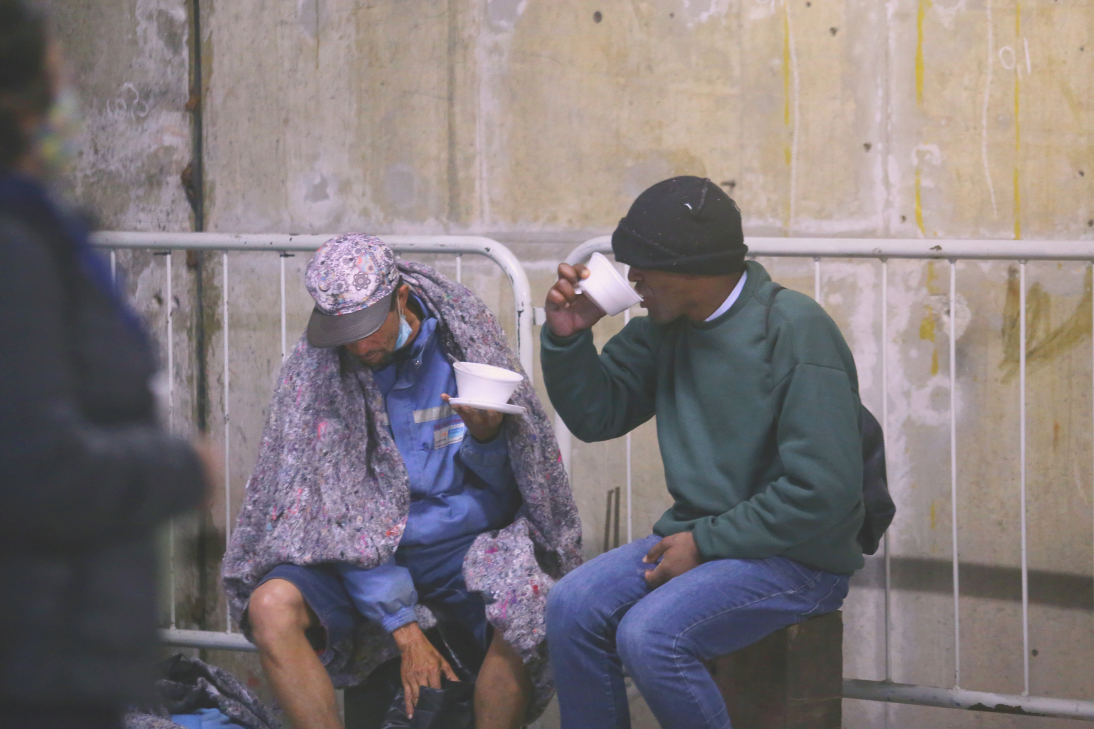 Fotografia contém dois moradores de rua tomando sopa de um recipiente de isopor. Eles se encontram dentro de estação metrô. Eles estão agasalhados e um deles está com um cobertor em volta dos ombros.