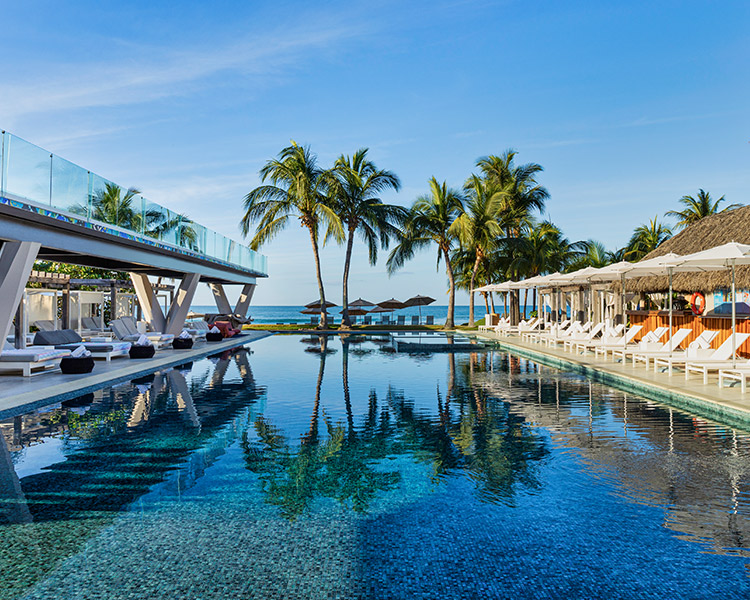 Luxurious resort pool with crystal-clear water, lined with palm trees and sun loungers under thatched umbrellas, with a clear blue sky in the background.