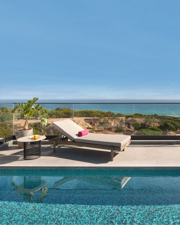 A serene poolside setting with a single lounge chair overlooking a sparkling ocean, framed by clear skies and a green landscape. a table with a drink and book adds a relaxing touch.