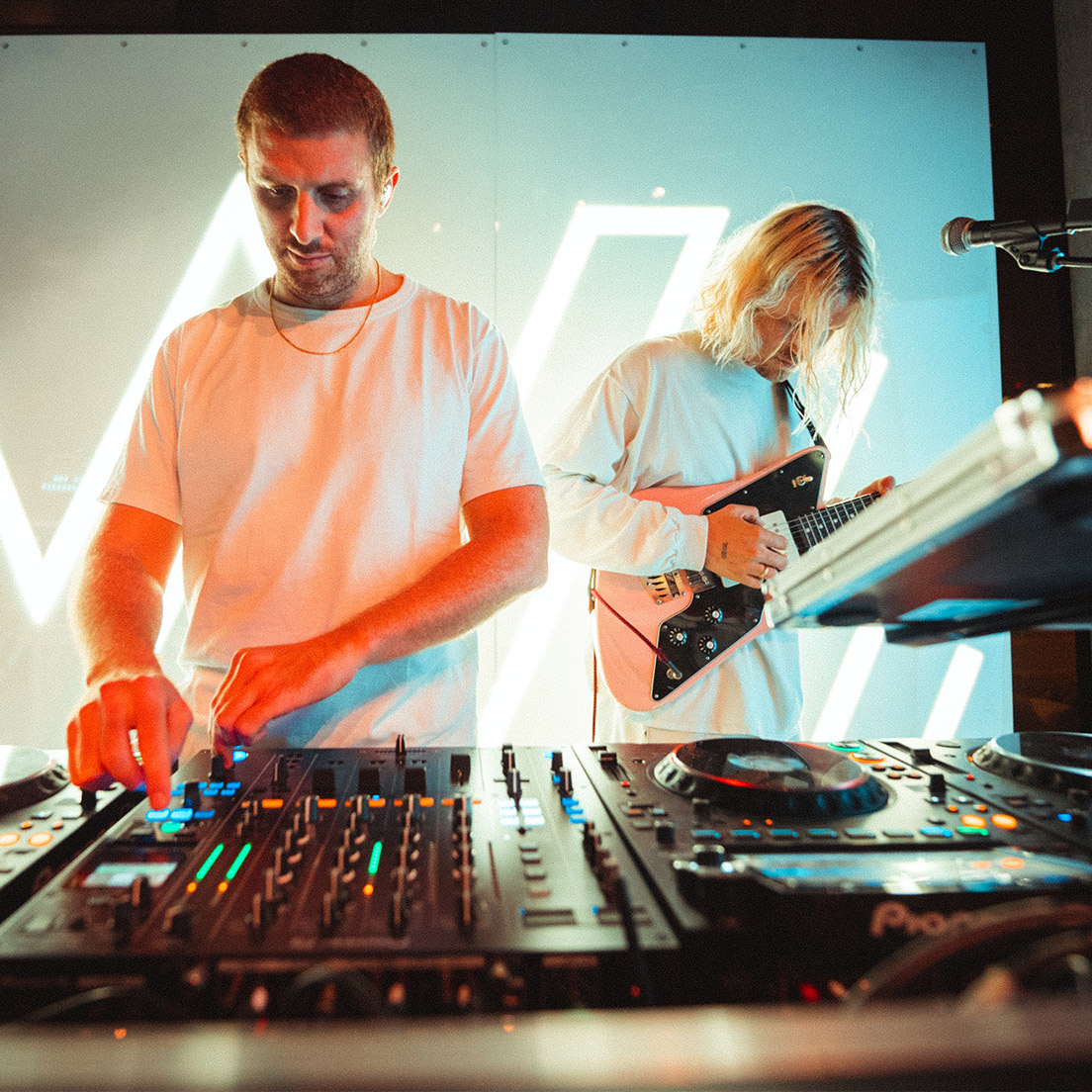 Two musicians on stage, one mixing on a dj console and the other playing an electric guitar, with vibrant stage lighting and electronic displays in the background.