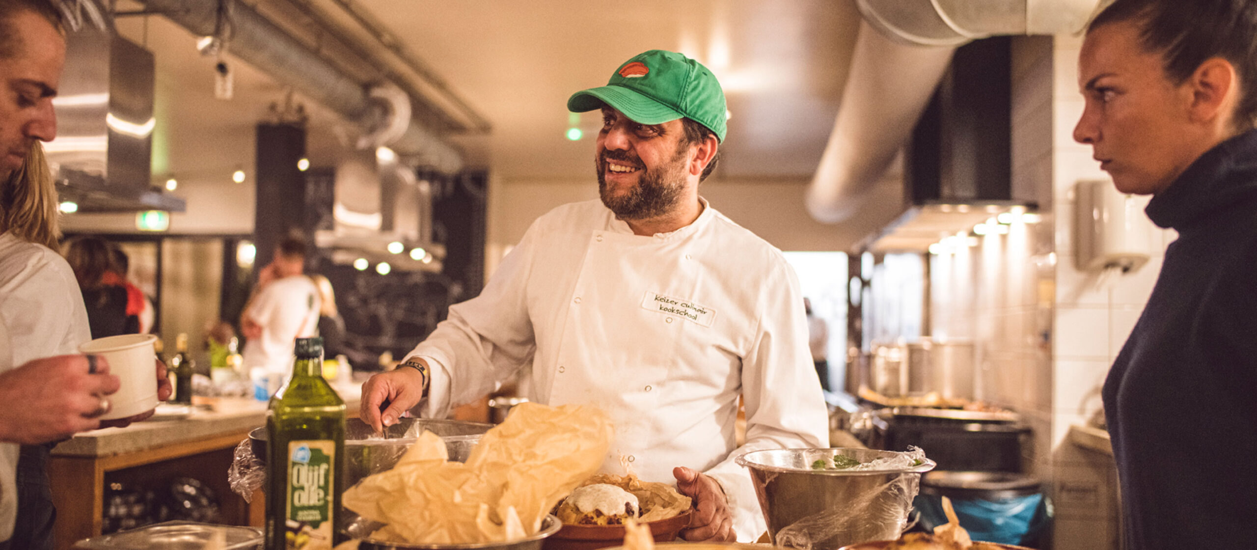 A chef wearing a white coat and green cap is smiling and talking to two people in a bustling kitchen. The counter is filled with various food items and ingredients. One person holds a bowl and another appears engaged in conversation. The atmosphere is lively and warm.