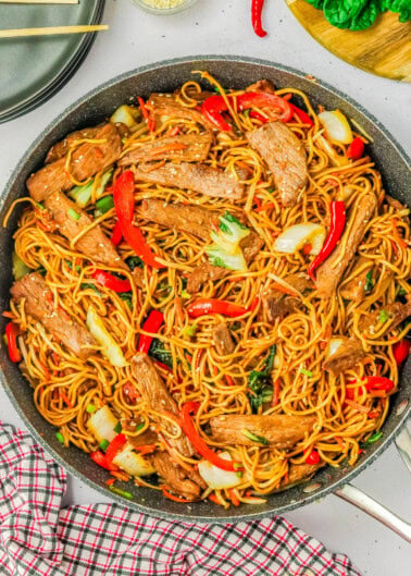 A pan of stir-fried noodles with slices of beef, red bell peppers, bok choy, and green onions. A red checkered cloth is underneath the pan. Plates and ingredients are visible in the background.