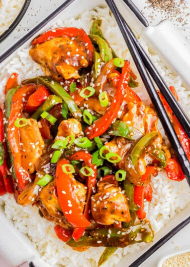 A close-up of a plate of stir-fry with chicken, bell peppers, and green onions served over white rice. Chopsticks rest on the plate.