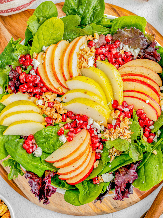 A fresh salad with sliced apples and pears, pomegranate seeds, walnuts, and crumbled cheese on a bed of mixed greens.